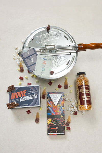 The LUX BOX co. Movie Night box features a stovetop popcorn popper, popcorn kernels, assorted sweet snacks, a Movie Charades game, and a movie-themed chocolate bar—all arranged for the ultimate Snack Delights experience.