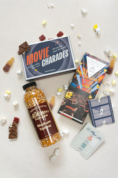Flat lay of LUX BOX co.’s Movie Night set: includes a Movie Charades game, popcorn jar, chocolate bar, candy, film strip coaster, tickets, and scattered popcorn—perfect for enjoying under the screen lights.