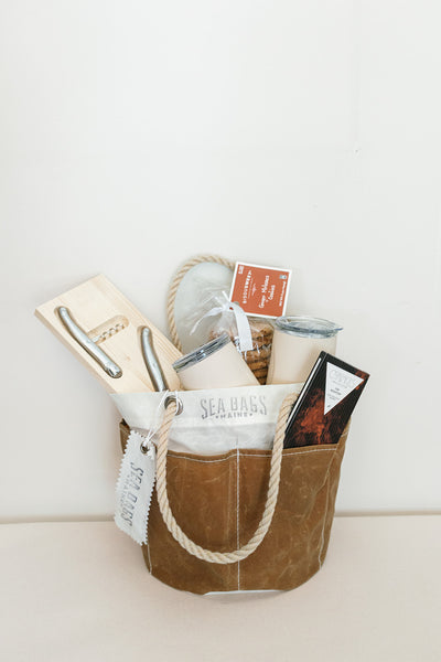 The Cocktails on the Trail gift by LUX BOX co. features a brown Sea Bags bucket tote with rope handles holding a wood cheese slicer, candle, chocolate bar, tin can, cocktail bottle, and notepad. The Sea Bags Maine label is visible.