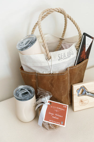 The Cocktails on the Trail gift set by LUX BOX co. features a brown Sea Bags bucket tote with rope handles holding a white tumbler, notebook, and pouch. In front are another tumbler for cocktails, ginger molasses cookies, and a wooden bottle opener.