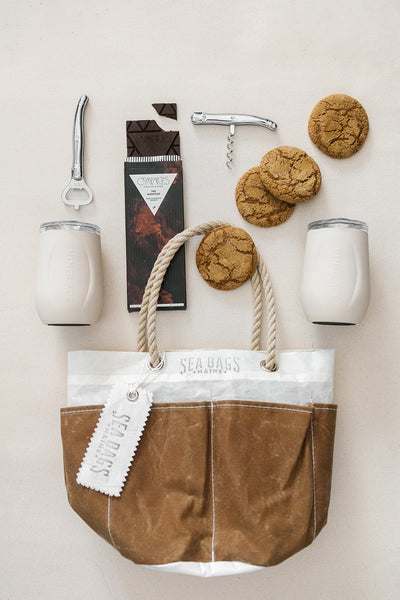 The Cocktails on the Trail gift by LUX BOX co. features a brown and white Sea Bags bucket tote with rope handles, two tumblers, a bottle opener, a corkscrew, a chocolate bar, three cookies, and a tag—ideal for cocktails on the go.