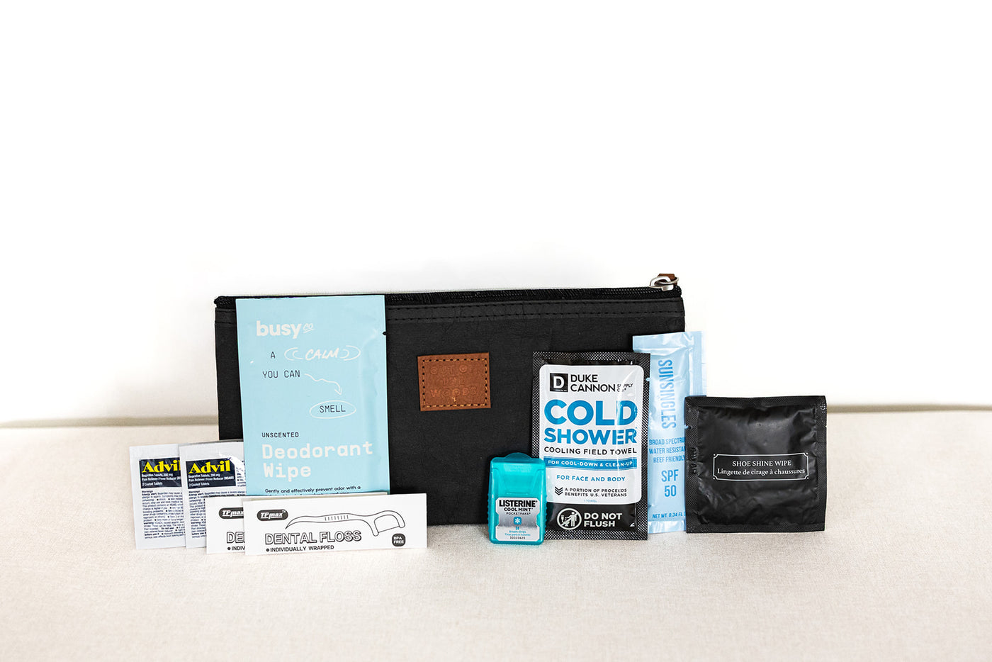 The LUX BOX co. Amenities on the Go - For Him is a black zippered pouch featuring travel-size essentials like deodorant wipes, cold shower towels, Advil, dental floss, mouthwash, and a compact black pack.