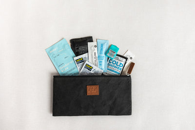 The LUX BOX co. Amenities on the Go - For Him is a black zippered pouch resting on a white surface, filled with travel-sized essentials like wipes, deodorant, lip balm, and cleansing packets.