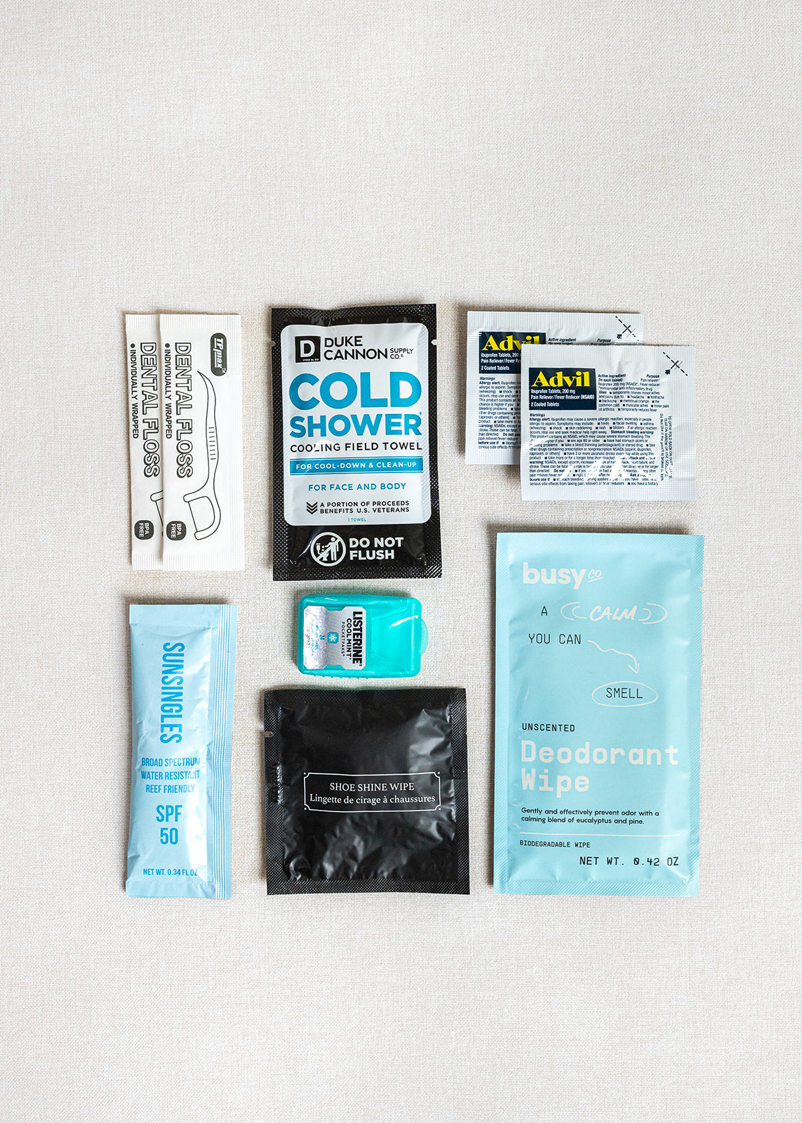 A flat lay of LUX BOX co.'s Amenities on the Go - For Him shows travel essentials like a cooling towel, Advil packets, dental floss, earplugs, sunscreen, shoe shine and deodorant wipes, and a mint on a light background.