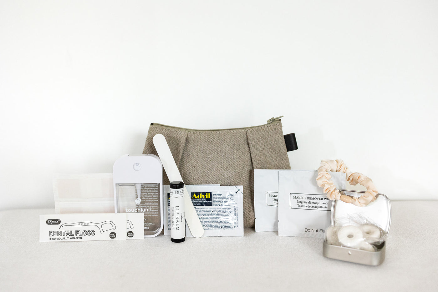 The LUX BOX co. Amenities on the Go – For Her kit includes a beige toiletry bag with dental floss, hand sanitizer, lip balm, Advil, makeup remover wipes, scrunchie, cotton swabs in a tin, nail file, and tissues arranged neatly.
