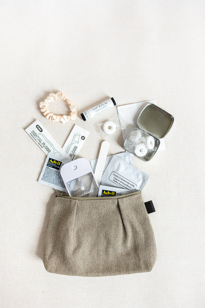 The LUX BOX co. Amenities on the Go - For Her pouch features a beige zippered bag with essentials like a scrunchie, lip balm, breath mints, nail file, bandages, floss picks, and wipes arranged on a light background.