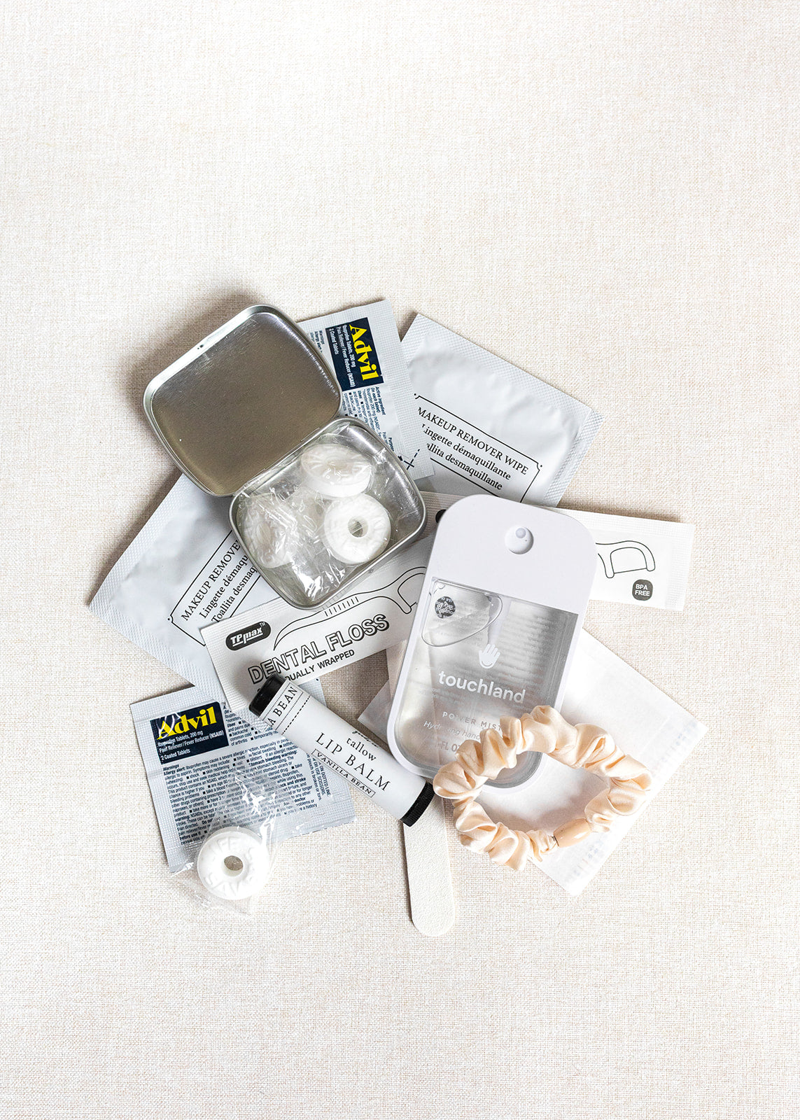 A flat lay of LUX BOX co.’s Amenities on the Go - For Her kit, featuring mints, dental floss, hand sanitizer, a nail file, satin scrunchie, lip balm packet, and sanitizing wipes arranged on a light surface.