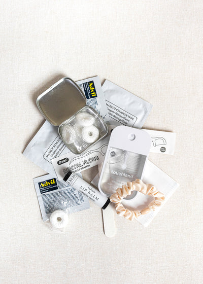 A flat lay of LUX BOX co.’s Amenities on the Go - For Her kit, featuring mints, dental floss, hand sanitizer, a nail file, satin scrunchie, lip balm packet, and sanitizing wipes arranged on a light surface.