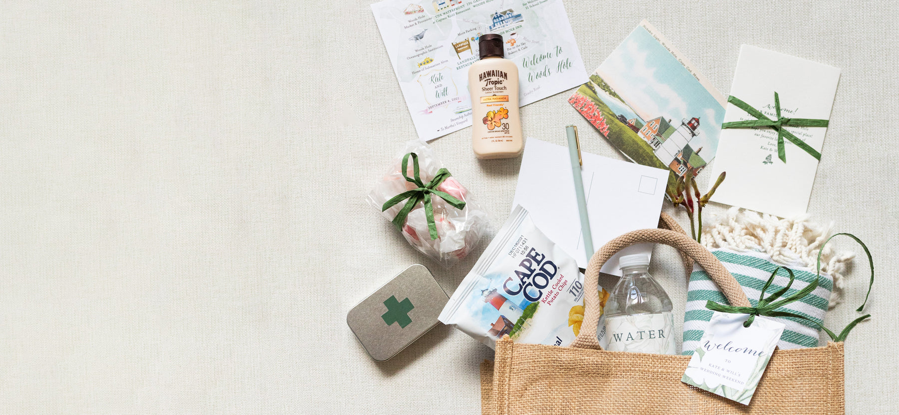 Open tote bag with various items from The Lux Box Co: bottled water, snack bag, small tin, wrapped soap, lotion, postcards, calendar sheet, and ribbon-tied envelope—all arranged on a light-colored surface. Perfect for corporate gifts or personal surprises.