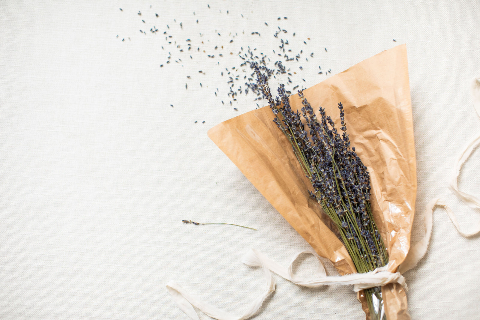 A bundle of dried lavender wrapped in brown paper lies on a white fabric surface with scattered buds around, perfect as part of The Lux Box Co's exquisite collection for Gifts for Social Events and Corporate Gifting.