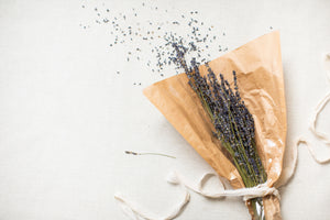 A bundle of dried lavender wrapped in brown paper lies on a white fabric surface with scattered buds around, perfect as part of The Lux Box Co's exquisite collection for Gifts for Social Events and Corporate Gifting.