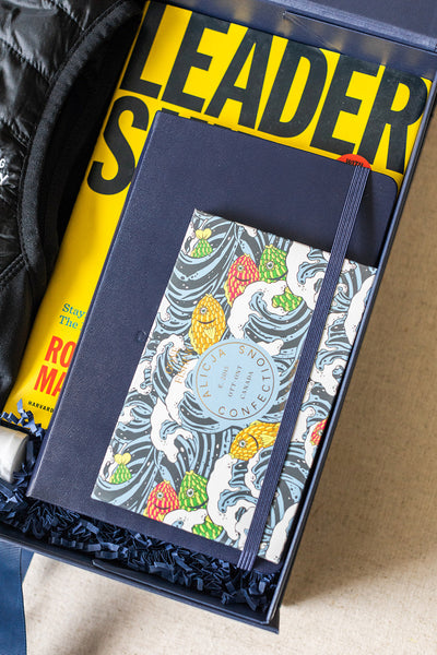 A gift box perfect for corporate gifting, featuring a yellow book titled "Leader S...", a blue notebook, and a colorfully illustrated book about confections.