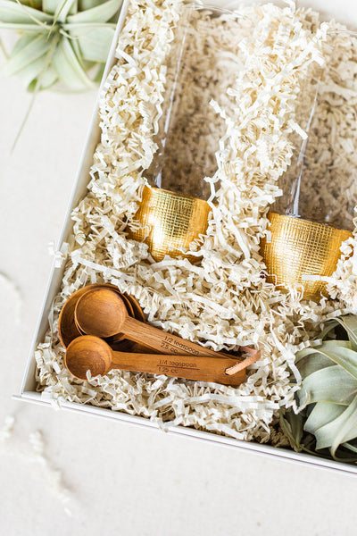 Discover a sophisticated gift box by The Lux Box Co, featuring two gold-rimmed glasses and a set of wooden measuring spoons nestled in white crinkle paper, accented by lush green plants. Perfect for corporate gifting or any special occasion that demands elegance.