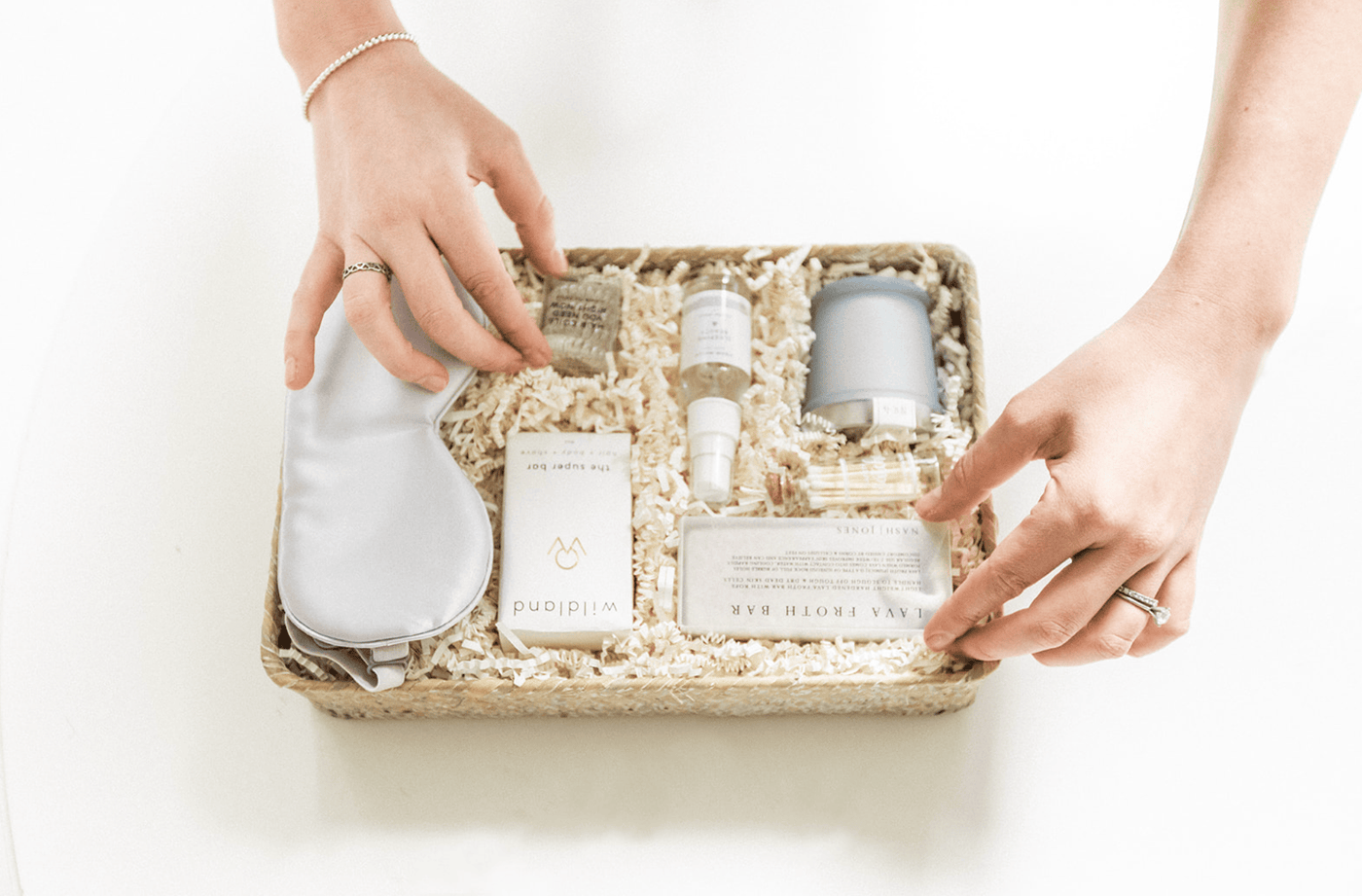 A person arranges a gift basket containing skincare products, an eye mask, a candle, and a soap bar, surrounded by shredded paper filler.