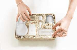 A person arranges a gift basket containing skincare products, an eye mask, a candle, and a soap bar, surrounded by shredded paper filler.