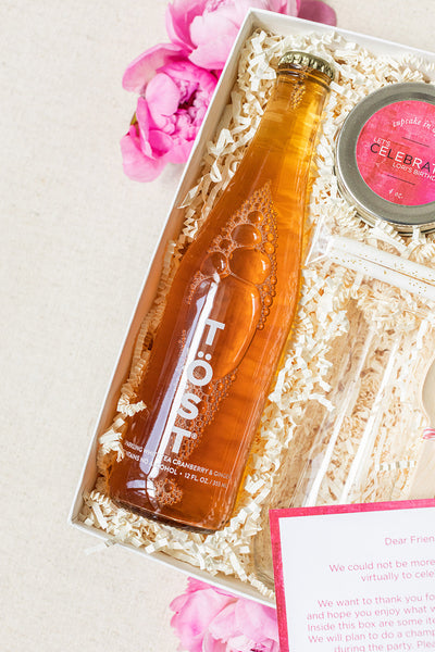 Gift box ideal for corporate gifting, featuring a bottle labeled "TOST," a round tin, and a heartfelt note on decorative shredded paper, surrounded by pink flowers. Perfect for adding elegance to social events.