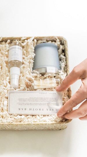 A hand reaches into a basket from The Lux Box Co, filled with beauty products and paper shreds—a spray bottle, candle, and bar of soap—perfect gifts for social events.