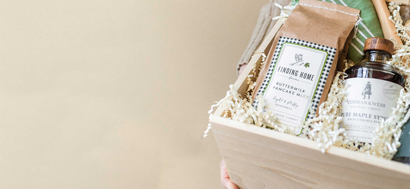 Wooden basket containing Finding Home Farms Buttermilk Pancake Mix, Crown Maple Syrup, and shredded paper filling.