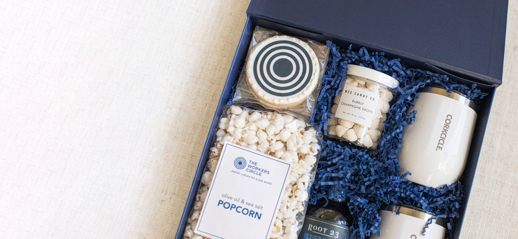Gift box containing popcorn, a decorative cookie, spiced peanuts, two insulated cups, and root 23 syrups, arranged with blue crinkle paper.