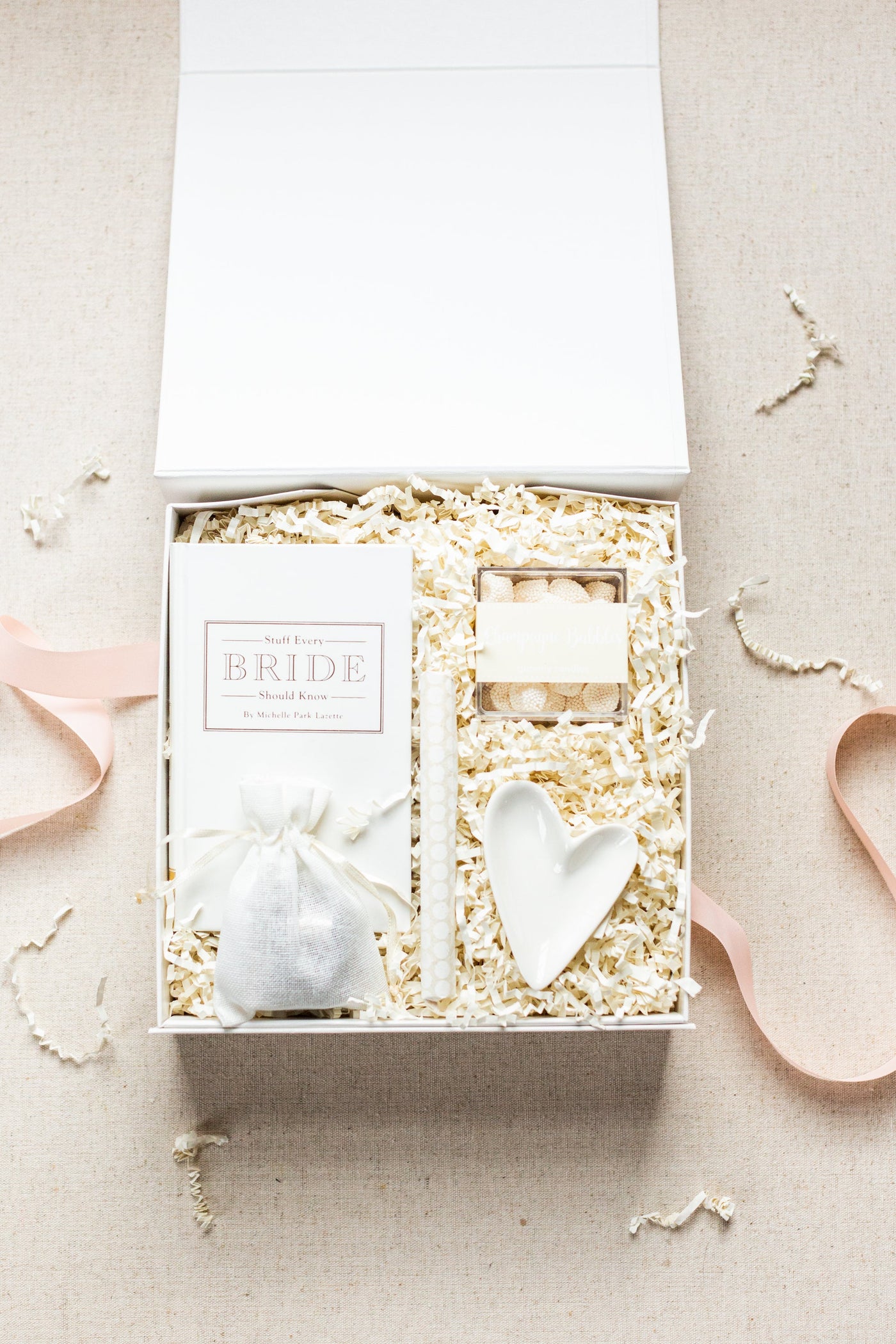 The LUX BOX Engagement Gift Box includes a book titled BRIDE, a candle, soap bar, heart-shaped dish, and drawstring pouch, all set in shredded paper. Its perfect for the wedding planners engagement gift and features pink ribbon accents against a beige background.