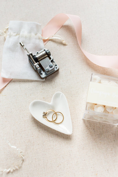 The LUX BOX Engagement Gift Box is an elegant choice, featuring a charming music box, two gold rings on a heart-shaped dish, a white pouch, and sugar cubes tied with a pink ribbon on beige cloth.