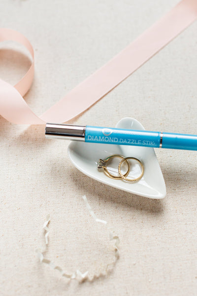A blue Diamond Dazzle Stik and two rings from the LUX BOX Engagement Gift Box sit on a heart-shaped dish, ideal for an engagement gift, with a pink ribbon and confetti nearby on a beige surface.