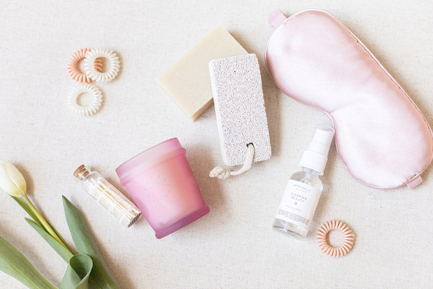 A flat lay of LUX BOXs Spa Gift Box - Blush includes a silk sleep mask, spray bottle, pink candle, pumice stone, bar soap, hair ties, matches, and tulip on a beige background—perfect for curating your own Beauty Sleep gift box.
