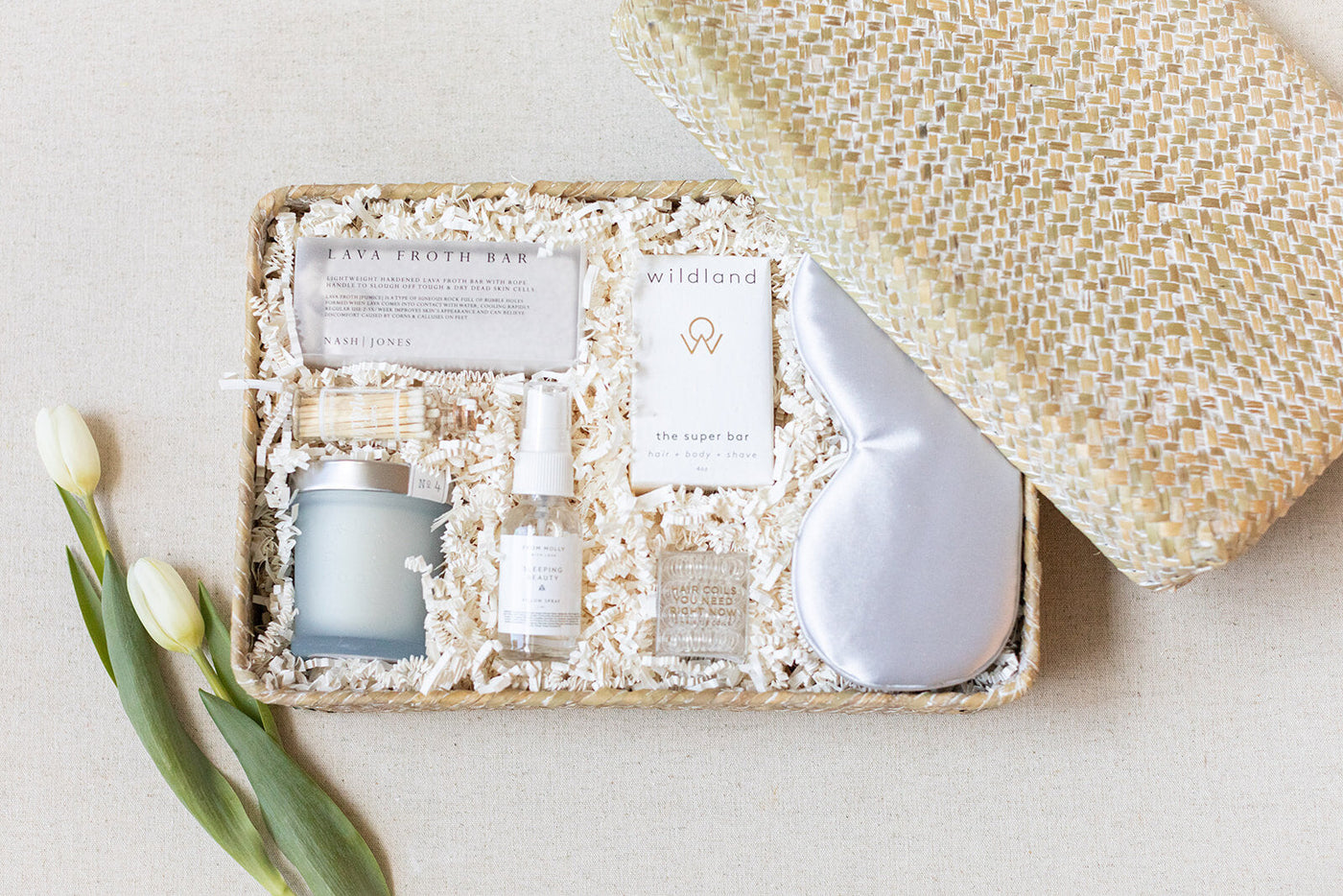 The LUX BOX Spa Gift Box - Grey features a bar soap, candle, spray, silk sleep mask, and informative pamphlets on shredded paper. Perfect for enhancing your self-care routine. Elegantly complete with two white tulips beside the open lid.
