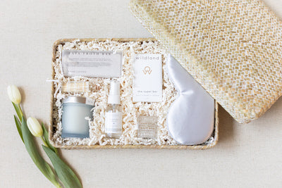 The LUX BOX Spa Gift Box - Grey features a bar soap, candle, spray, silk sleep mask, and informative pamphlets on shredded paper. Perfect for enhancing your self-care routine. Elegantly complete with two white tulips beside the open lid.