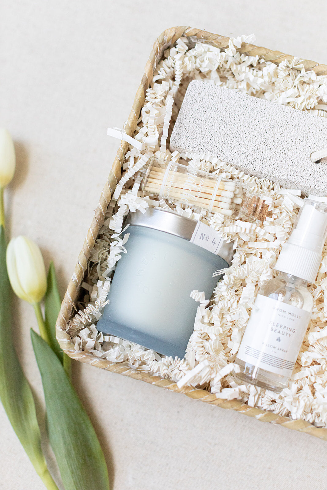 The LUX BOX Spa Gift Box - Grey includes a candle, spray bottle, matches, and a pumice stone in shredded paper. Enhance your self-care routine with this collection. Two white tulips and a silk sleep mask add serenity to the light background.