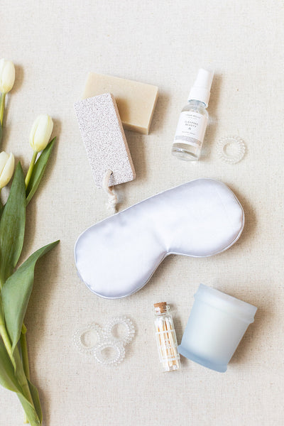 Items from the LUX BOX Spa Gift Box - Grey on a beige surface include a silk sleep mask, soap, spray bottle, candle, small bottle, hair ties, and pumice stone. White tulips are on the left.
