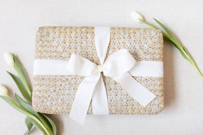 The LUX BOX Spa Gift Box - Grey, wrapped in woven fabric with a white ribbon and bow, sits on a light background with three white tulips nearby, offering tranquility and beauty sleep bliss.