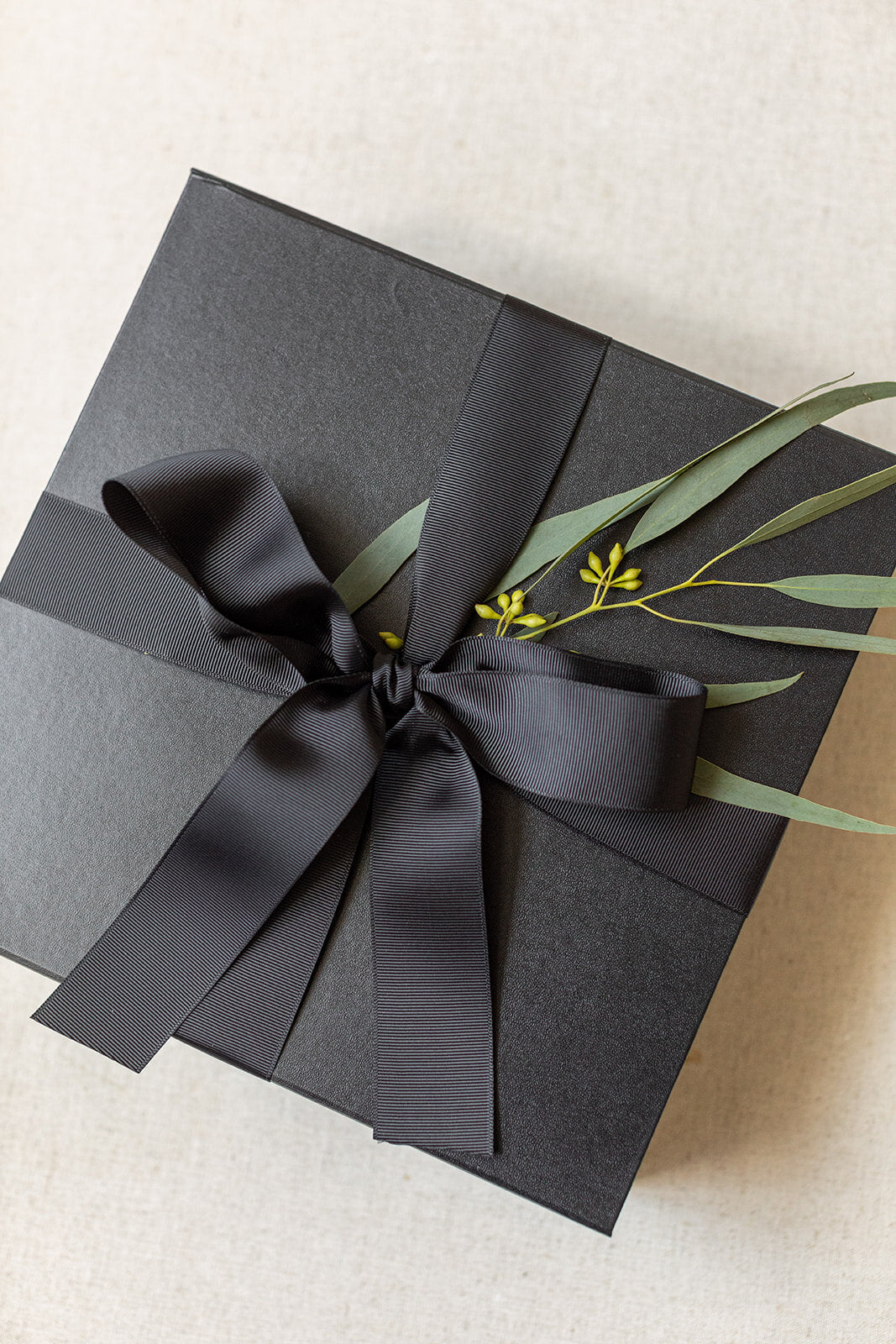 A sleek LUX BOXs Corporate Suite Gift Box, adorned with a black ribbon bow and green foliage, sits elegantly on a light background.