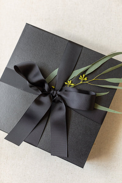 A sleek LUX BOXs Corporate Suite Gift Box, adorned with a black ribbon bow and green foliage, sits elegantly on a light background.