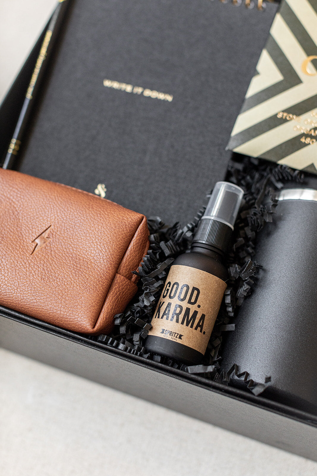 The Corporate Suite Gift Box by LUX BOX includes a brown wallet, black notebook, striped envelope on black shredded paper, and Good Karma focus-boosting spray for enhanced clarity.