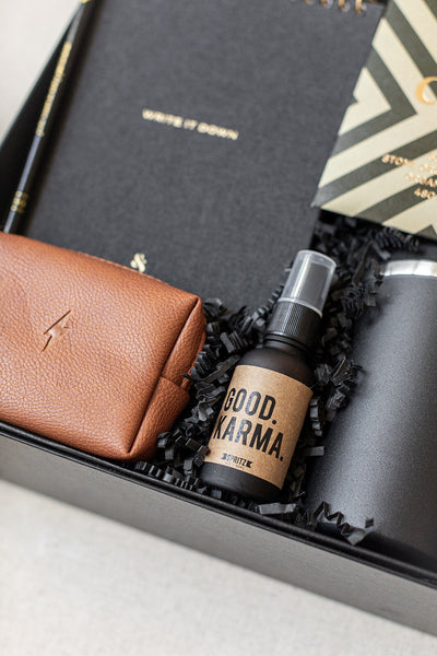 The Corporate Suite Gift Box by LUX BOX includes a brown wallet, black notebook, striped envelope on black shredded paper, and Good Karma focus-boosting spray for enhanced clarity.