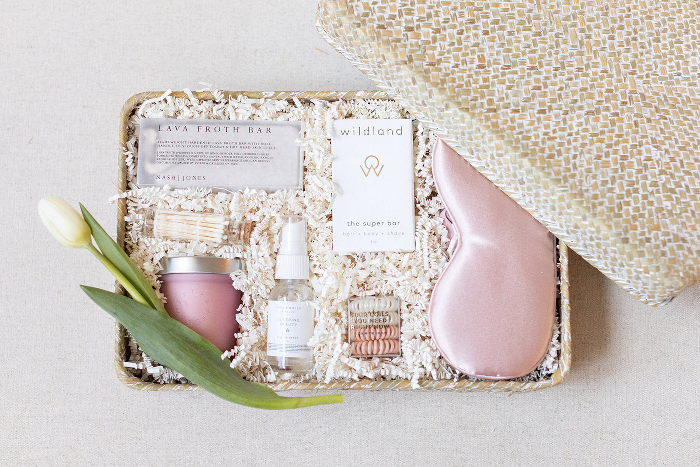 Introducing the LUX BOX Spa Gift Box - Blush: a luxurious set featuring skincare products, soap bars, a pink silk sleep mask, matches, a candle, and a white tulip on shredded packing material—the ultimate secret to rejuvenation.
