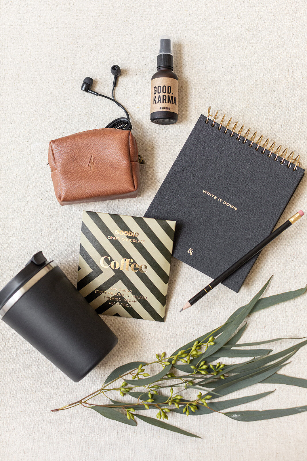 The Corporate Suite Gift Box by LUX BOX includes a curated selection on a beige surface: earphones, brown pouch, Focus Boosting Spray, notebook with pencil, black tumbler, coffee packet, and eucalyptus leaves with small yellow flowers. Perfect for staying energized and centered.