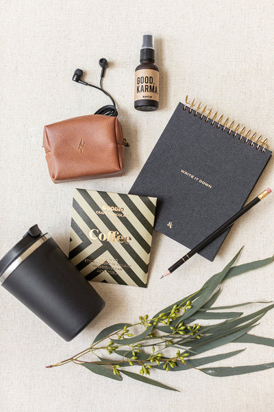 The Corporate Suite Gift Box by LUX BOX includes a curated selection on a beige surface: earphones, brown pouch, Focus Boosting Spray, notebook with pencil, black tumbler, coffee packet, and eucalyptus leaves with small yellow flowers. Perfect for staying energized and centered.