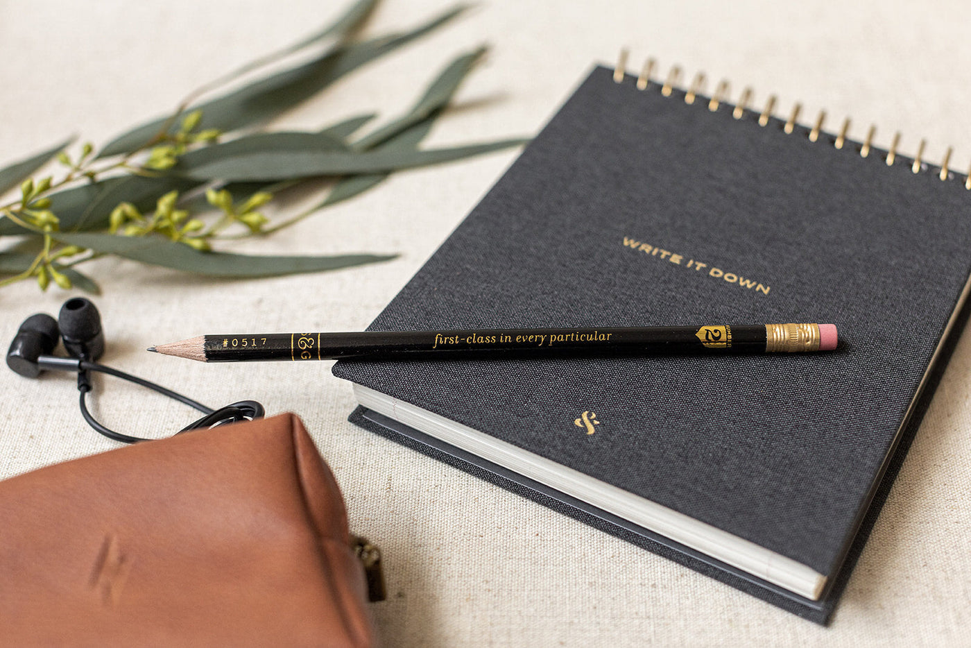 On a beige surface with green leaves, The Corporate Suite Gift Box by LUX BOX includes a spiral notebook saying Write It Down, a pencil, brown pouch, and black earphones accompanying the Tech Kit.