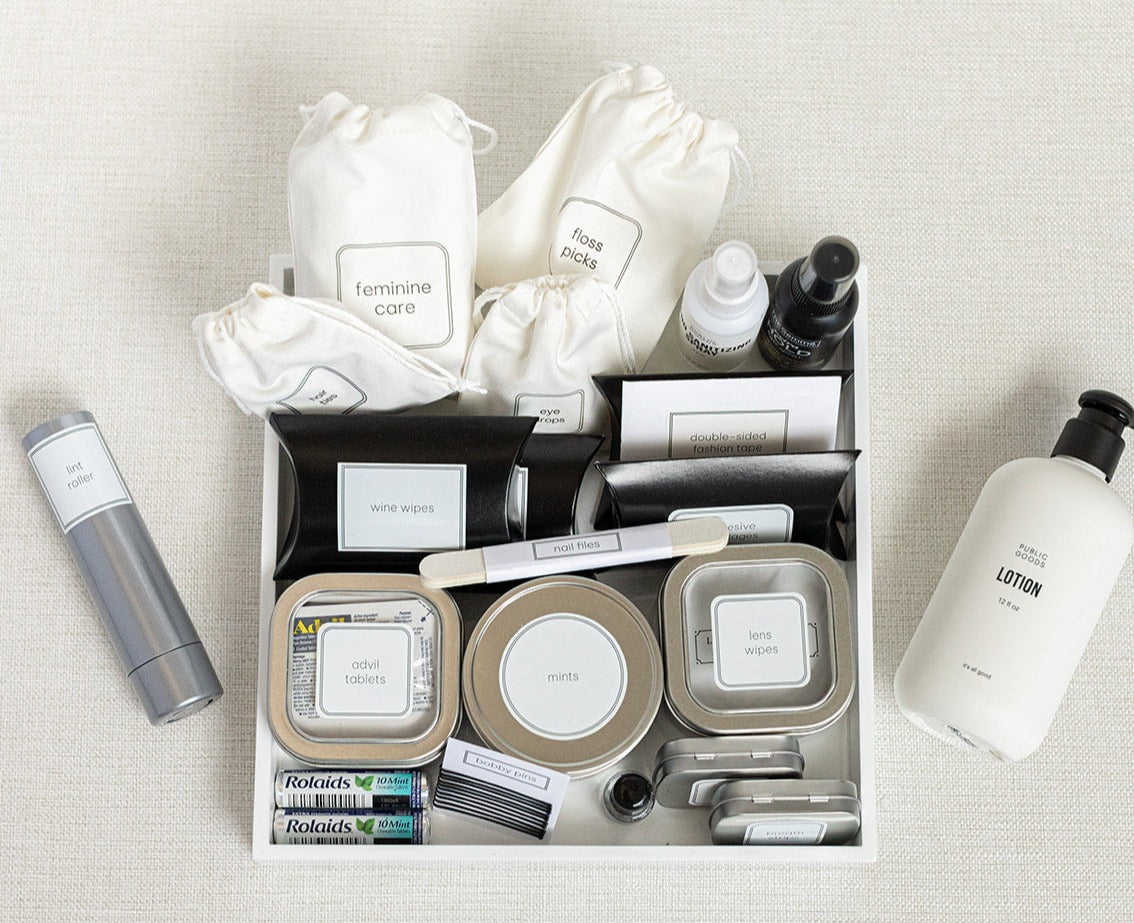The LUX BOXs Bathroom Amenities Tray - THE HERS offers a neatly arranged selection of labeled travel essentials, including feminine care, fashion tape, stain removal wipes, mints, lotion, and medications on a textured surface.