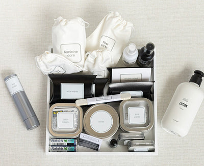 The LUX BOXs Bathroom Amenities Tray - THE HERS offers a neatly arranged selection of labeled travel essentials, including feminine care, fashion tape, stain removal wipes, mints, lotion, and medications on a textured surface.