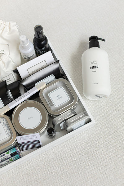 The HERS Bathroom Amenities Tray by LUX BOX displays toiletries like tins, spray bottles, stain removal wipes, and a toothbrush neatly arranged. A separate lotion bottle is gracefully placed on the side.