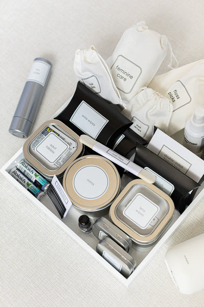 The LUX BOX Bathroom Amenities Tray - THE HERS neatly organizes travel essentials like stain removal wipes, feminine care products, mint tins, a razor, and hygiene products in labeled containers on a white surface.