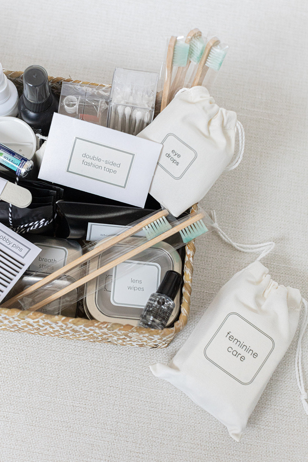 A basket teeming with essentials like toothbrushes, fashion tape, and wipes. Nearby, two drawstring bags labeled eye drops and feminine care complement the comprehensive LUX BOXs THE DELUXE Bathroom Amenities Tray.