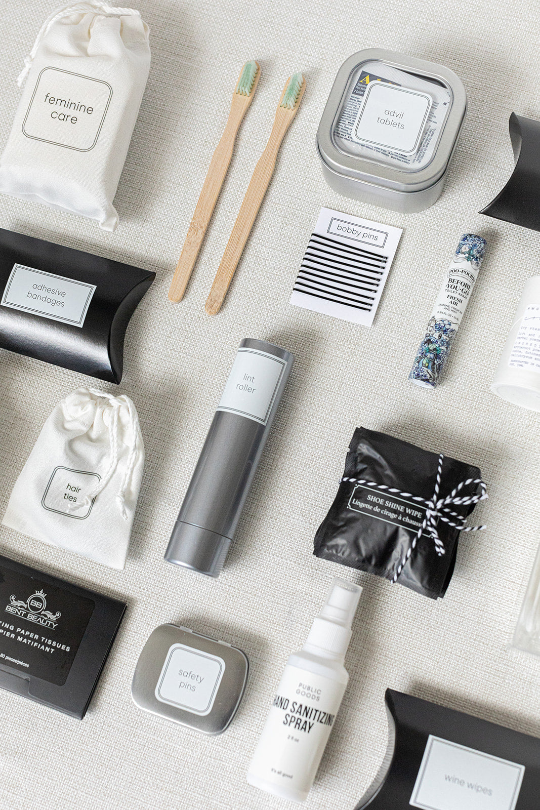 The LUX BOX Bathroom Amenities Tray - THE DELUXE offers a well-arranged selection of essentials like toothbrushes, sanitary spray, bobby pins, safety pins, lint roller, and feminine care products on a light fabric surface.