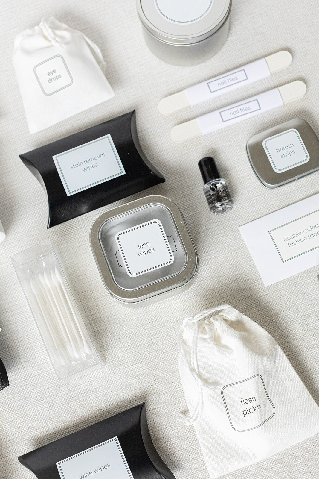 A flat lay features the LUX BOX Bathroom Amenities Tray, THE DELUXE, elegantly arranged on a light surface with tins, bottles, and pouches labeled for uses like lens wipes, floss picks, and nail files.
