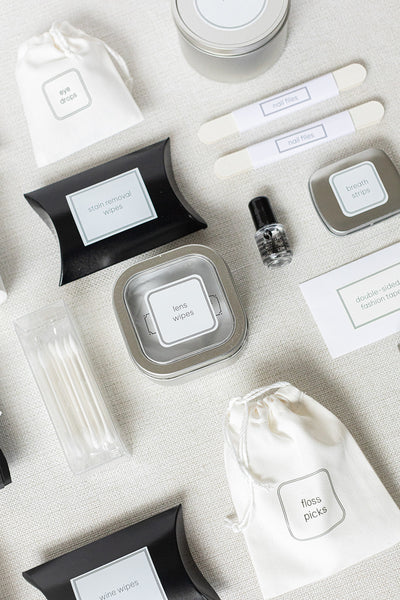 A flat lay features the LUX BOX Bathroom Amenities Tray, THE DELUXE, elegantly arranged on a light surface with tins, bottles, and pouches labeled for uses like lens wipes, floss picks, and nail files.